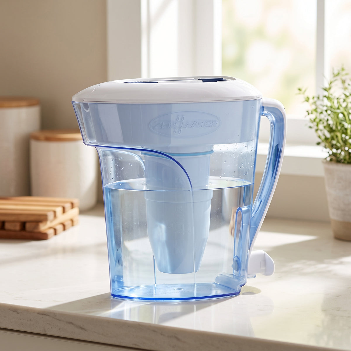 A ZeroWater water filter pitcher with a translucent light blue body and a white lid, sitting on a light marble kitchen countertop with a window and kitchen items blurred in the background.